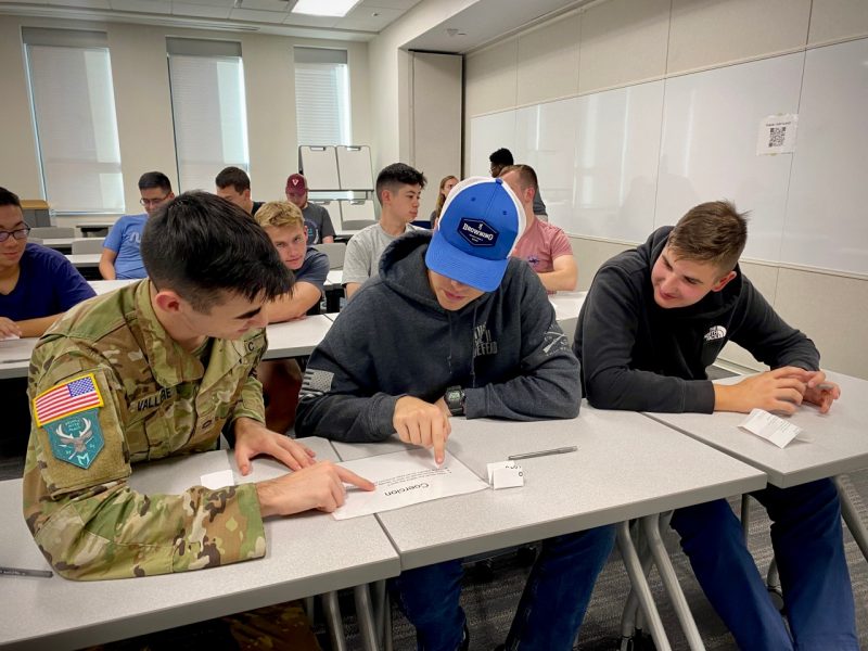 Three cadets view a sheet of paper on a desk. The classroom behind them is filled with other cadets in the class.