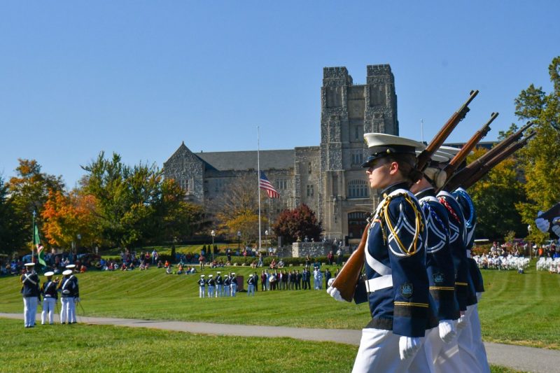 The front rank of a cadet formation marches onto the drillfield for a parade. Burruss in in the background and the cadets are in their dress uniforms holding rifles. The  first two cadets are women.