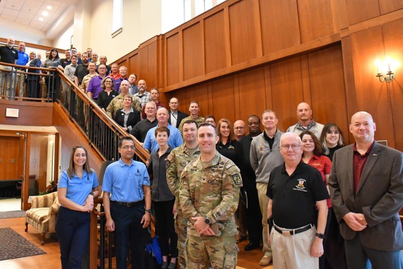 A large group of instructors fills a wooden stairway at the alumni center. They are all looking at the camera and smiling. 