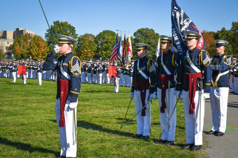 A cadet battalion commander in his dress uniforms salutes with his sword as cadets stand in the background saluting.