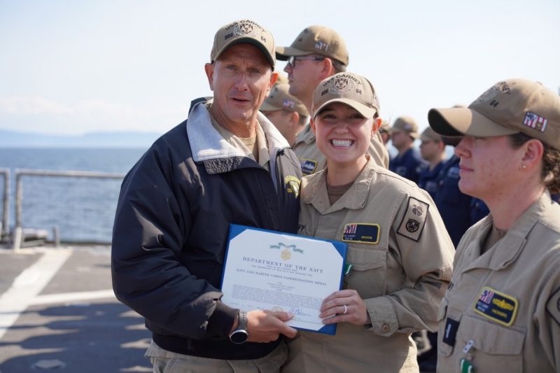 Robertson and Mason stand facing the camera holding a certificate. Mason is smiling.