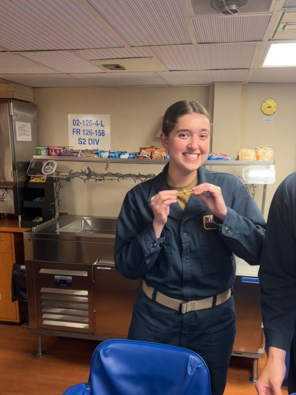 Mason stands on the ship in uniform holding a golden military pin. She is smiling.