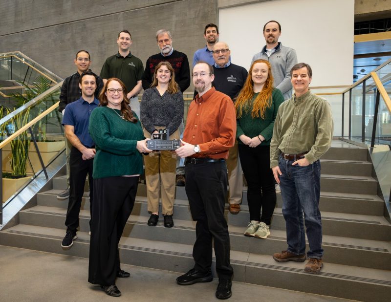 A group of people stand on stairs smiling at the camera while holding an award