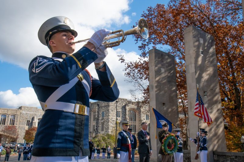 A cadet in dress uniform plays the bugle while a wreathe is laid in the background at the Pylons