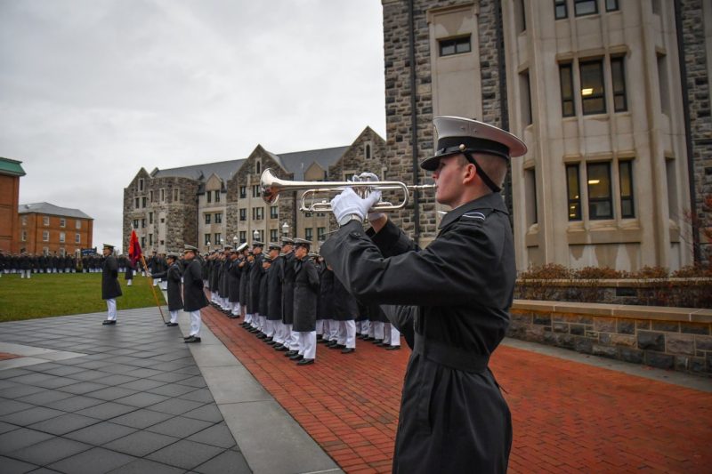 A cadet in uniform plays the bugle while cadets in the background stand in formation