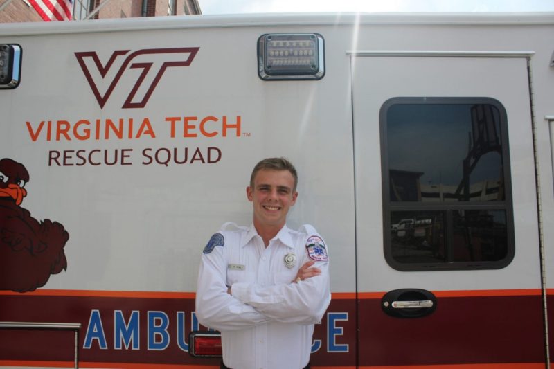 Feey stands in an EMT uniform smiling in front of an ambulance saying VT Rescue Squad