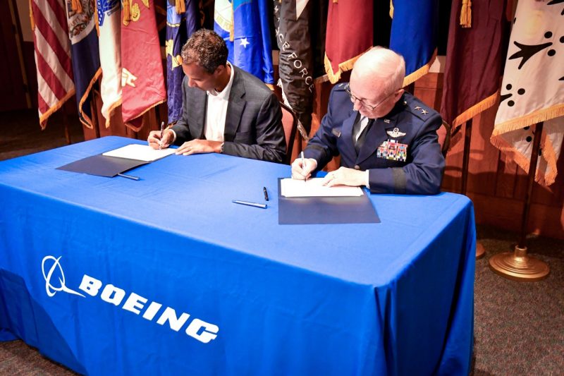 Allen and Fullhart sit at a skirted Boeing table with flags in the background 