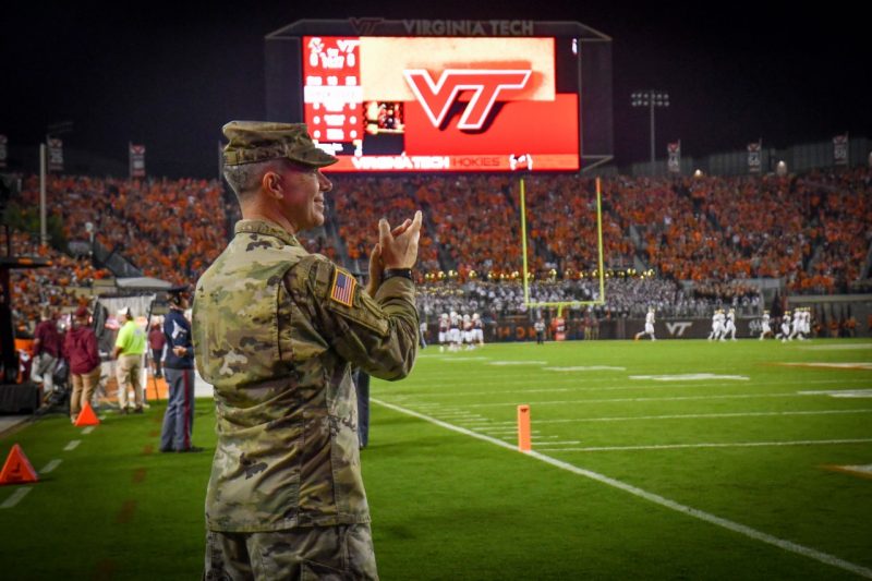Sgt. Maj. Combs claps and smiles while standing on the field for a night game at Lane Stadium. The scoreboard in the background shows an orange VT.