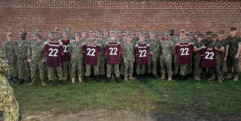 A large group of midshipmen stand together, some holding up maroon football jerseys with the number "22" on them.