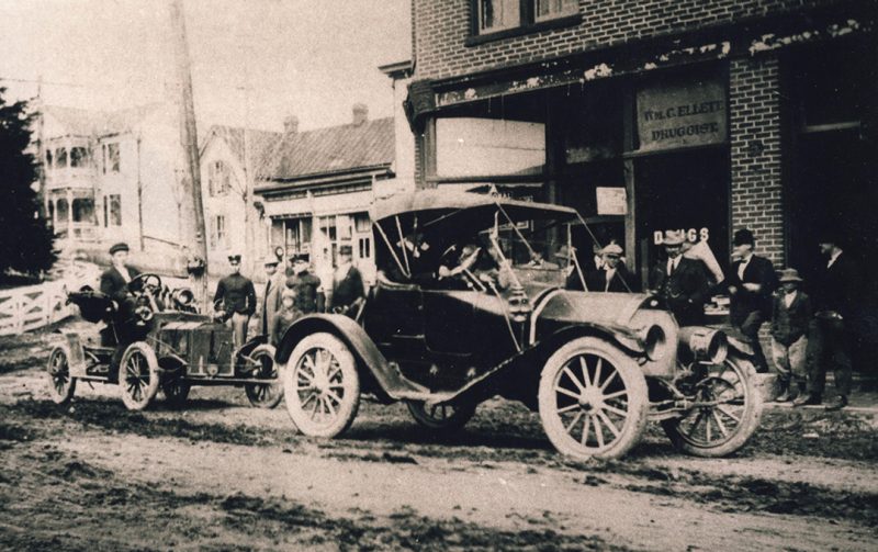 Two cars are parked in front of a store in a scene from the 1920s. 
