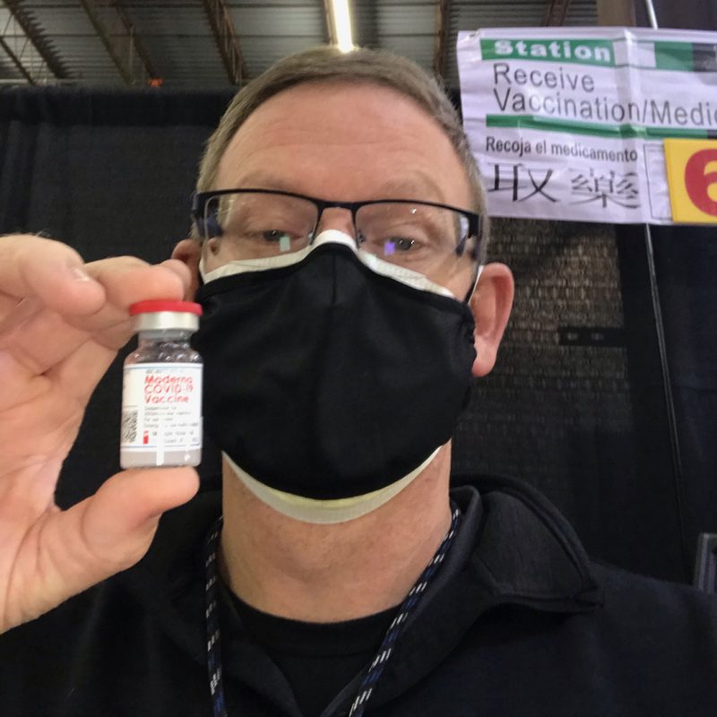Jeff Kircher E’94  wears a face mask and holds up a vial containing a COVID-19 vaccine.